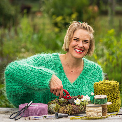 Lachende Frau beim Basteln eines Blumengestecks mit Gartenschere und Dekobändern im Freien